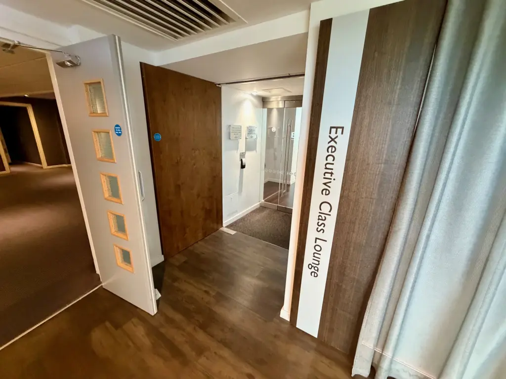 Entrance to an Executive Class Lounge with a tall sign reading 'Executive Class Lounge', a wood door, and glass elevator doors in the background.