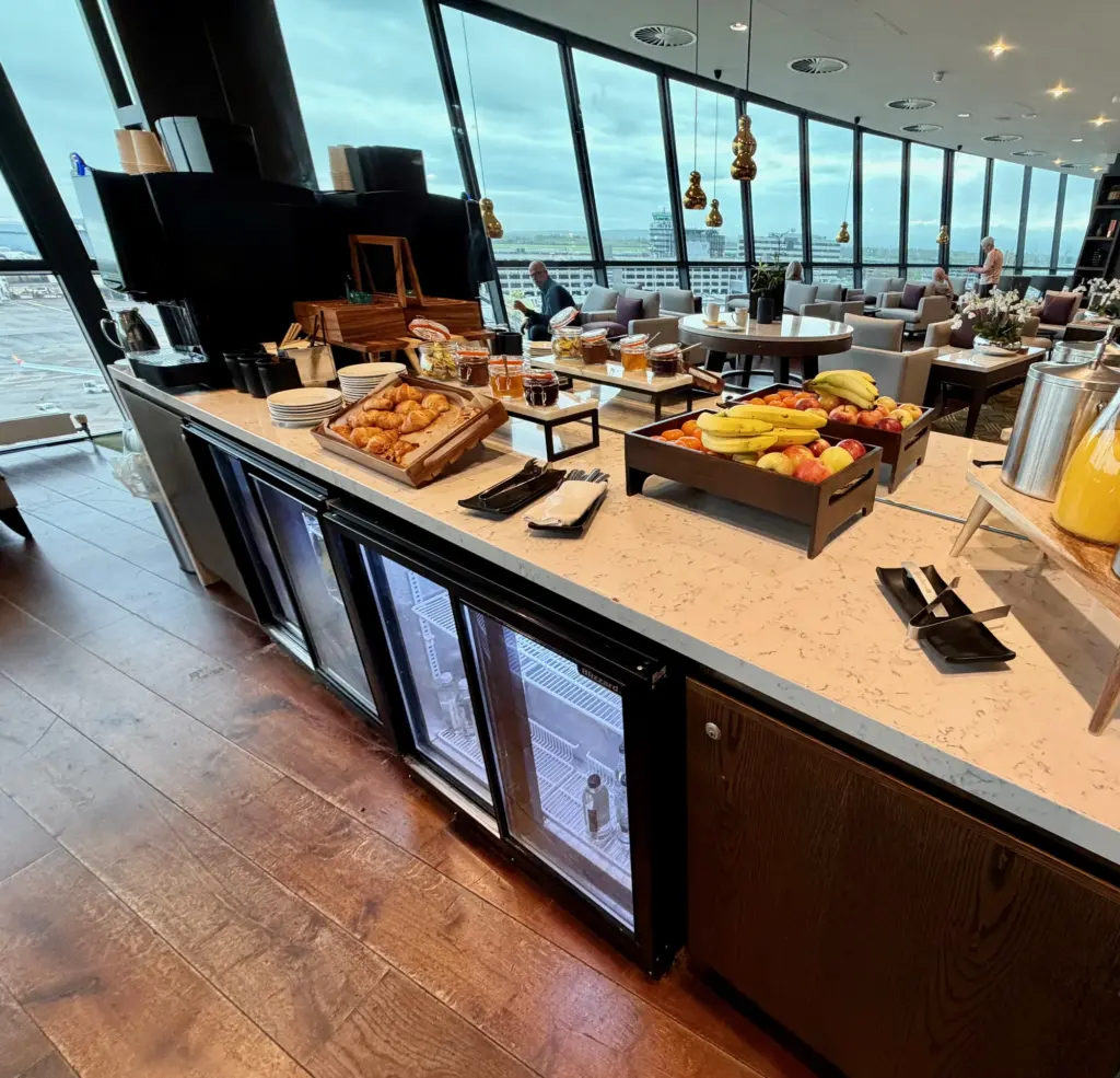 Buffet counter in a modern lounge with croissants, pastries, bowls of fruit, jars of jams, and juice carafes, set near large windows.
