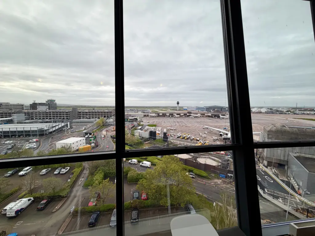 View of an airport apron from inside a terminal window: planes at gates, jet bridges, and distant terminal buildings under cloudy skies.
