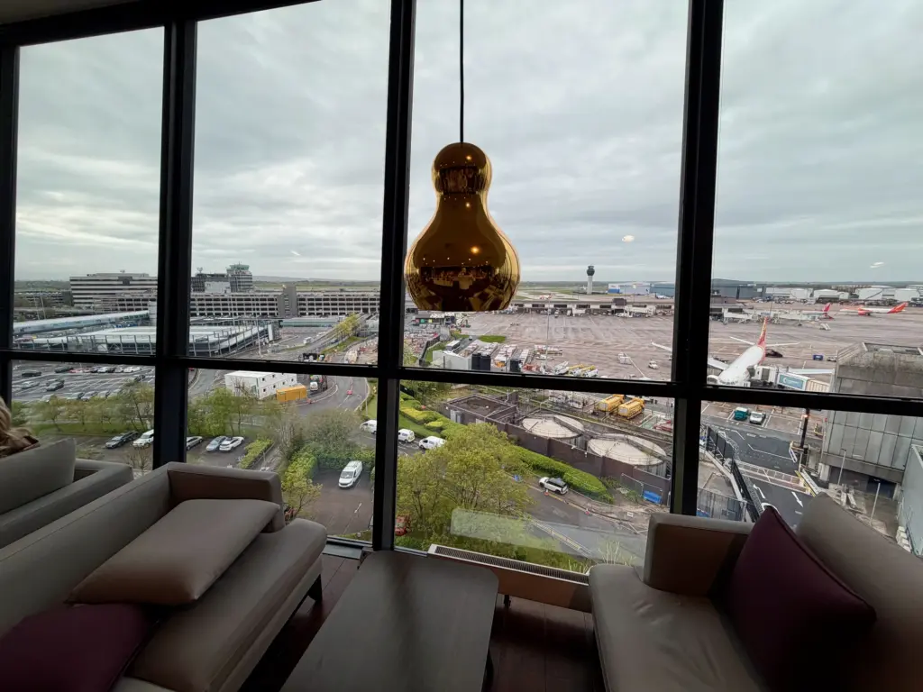 Lounge seating by a large window overlooking an airport tarmac with planes and a distant control tower; a gold pendant lamp hangs in foreground.