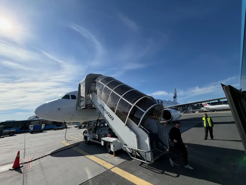 Disembarking our Finnair flight at a remote stand Airplane connected to a curved jet bridge at a sunny tarmac, Finnair branding visible, ground crew in high-visibility vests nearby.